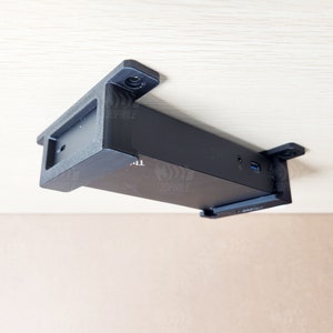 May include: A black device with a rectangular shape is mounted to the underside of a light wood surface. The device has a small black bracket on each end and a small black port on the side.