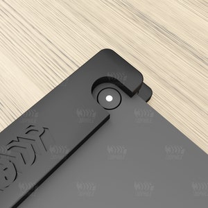 Puede incluir: Primer plano de una funda de tel&eacute;fono negra con un peque&ntilde;o c&iacute;rculo blanco en el centro. La funda tiene un borde elevado alrededor del objetivo de la c&aacute;mara y una superficie texturizada.