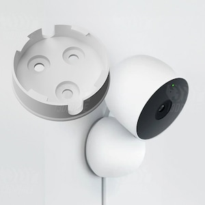 Puede incluir: Una cámara de seguridad blanca con una lente negra y una luz verde. La cámara está montada en un soporte de pared blanco. También se muestra un soporte de pared blanco separado.