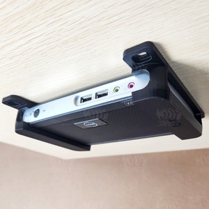 May include: A black computer with a silver top is mounted to the underside of a wooden surface using a black bracket. The computer has a Dell logo on the front.