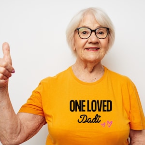 Könnte beinhalten: Eine Frau trägt ein gelbes T-Shirt mit dem Text "ONE LOVED Dadi" und zwei rosa Herzen. Sie lächelt und zeigt den Daumen nach oben.