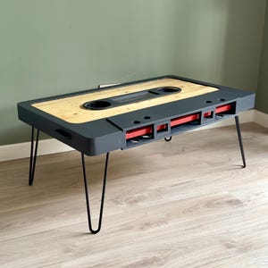Könnte beinhalten: Ein Couchtisch im Kassetten-Tape-Design. Er hat einen grauen Rahmen, eine helle Holzplatte und rote Akzente. Der Tisch steht auf schwarzen Haarnadelbeinen. Die Maße betragen ca. 120 cm Länge, 60 cm Breite und 40 cm Höhe, für einen Retro-Look.