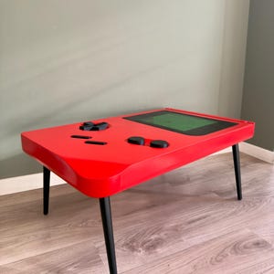 May include: A red coffee table designed to resemble a classic handheld video game console. The table features black control buttons and a screen, with black legs. The table is a unique piece of furniture.