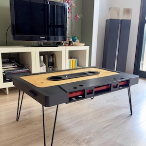 Puede incluir: Una original mesa de centro con forma de cinta de cassette. La mesa tiene un marco negro, una tapa de madera y detalles rojos. Se apoya en patas de horquilla negras, un mueble creativo.