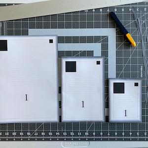 Può includere: Tre fogli di carta bianchi con quadrati neri nell'angolo in alto a sinistra e il numero "1" stampato al centro in basso. I fogli sono incorniciati da un bordo grigio e sono disposti in ordine decrescente di dimensione su un tappetino da taglio grigio.