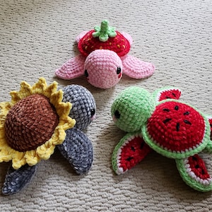 May include: Three crocheted turtle toys, each with a different fruit design. One is a pink strawberry turtle, one is a green watermelon turtle, and one is a gray sunflower turtle.
