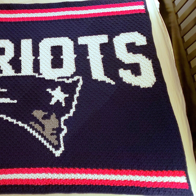 May include: A navy blue crocheted blanket featuring the word "PATRIOTS" in white yarn and a grey outline of the New England Patriots logo. The blanket has red and white stripes along the bottom edge.
