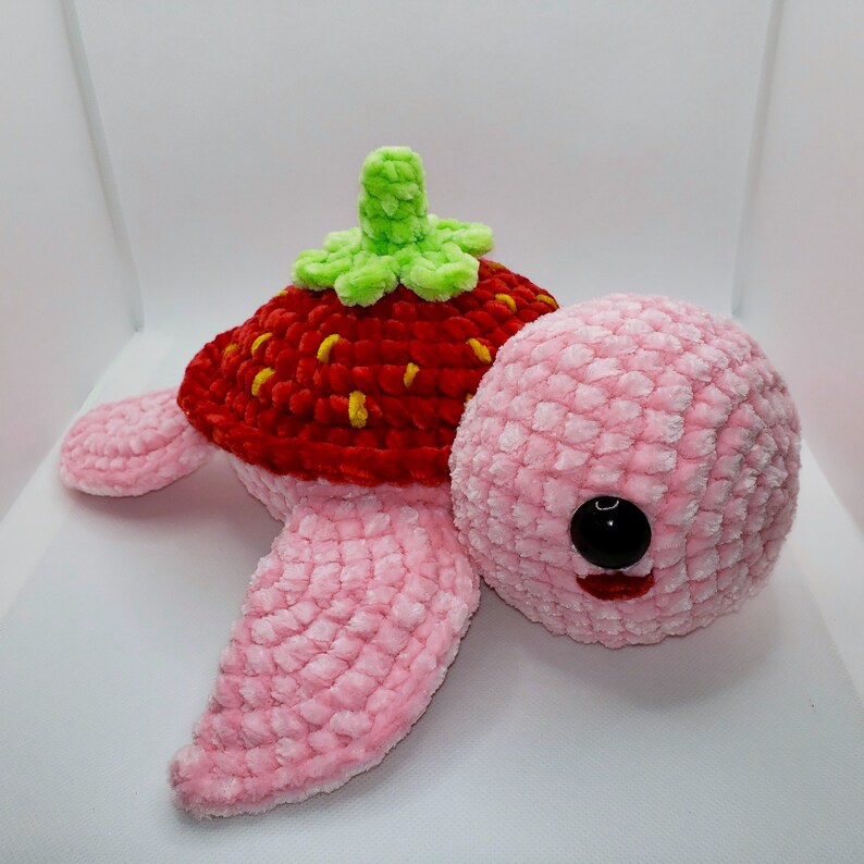 May include: A pink crocheted turtle with a red and yellow crocheted strawberry shell and a green crocheted leaf on top.