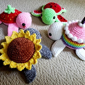 May include: Four crocheted turtle toys. One is pink with a strawberry on its back, one is green with a watermelon on its back, one is white with a rainbow and a pink frosting cake on its back, and one is gray with a yellow sunflower on its back.