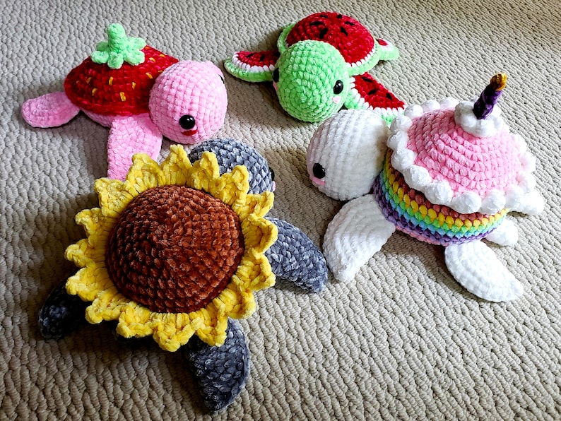 May include: Four crocheted turtle toys. One is pink with a strawberry on its back, one is green with a watermelon on its back, one is white with a rainbow and a pink frosting cake on its back, and one is gray with a yellow sunflower on its back.