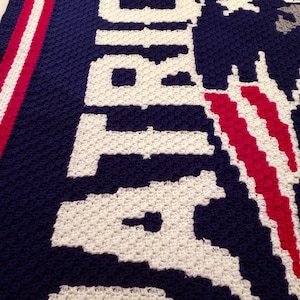 May include: Crocheted blanket featuring the word "Atlanta" in white yarn on a navy blue background. The blanket has red and white stripes on the left side.