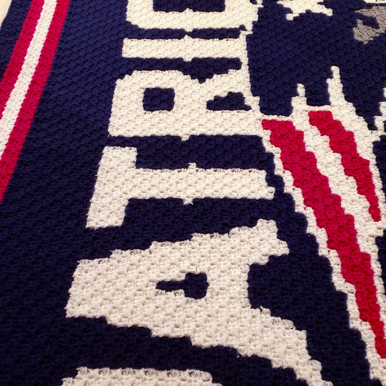 May include: Crocheted blanket featuring the word "Atlanta" in white yarn on a navy blue background. The blanket has red and white stripes on the left side.
