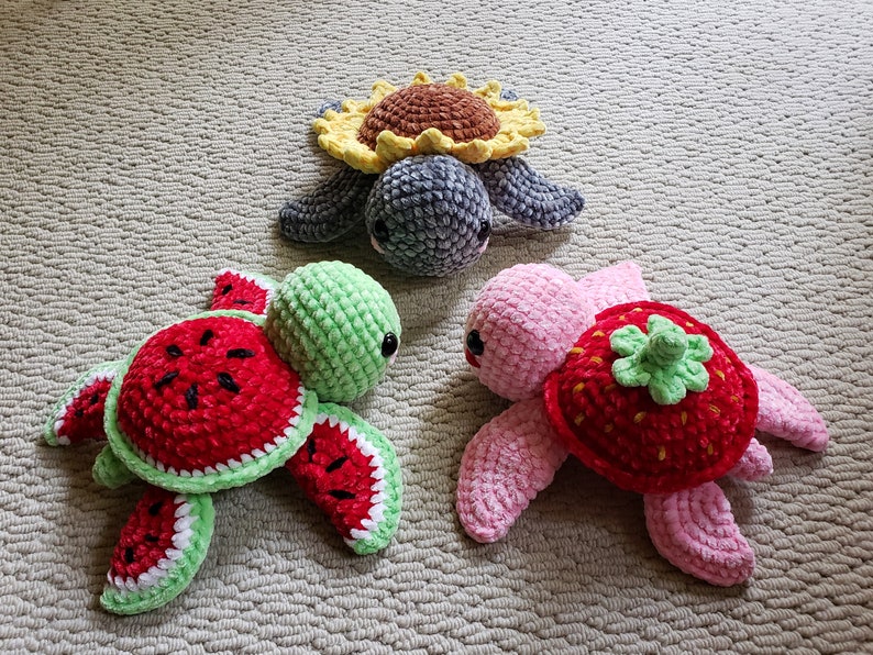 May include: Three crocheted turtle toys. One is green with a watermelon shell, one is pink with a strawberry shell, and one is gray with a sunflower shell.