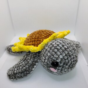 May include: A crocheted stuffed toy turtle with a yellow and brown sunflower on its back. The turtle has a gray body with a textured surface and a single black eye with a pink nose.