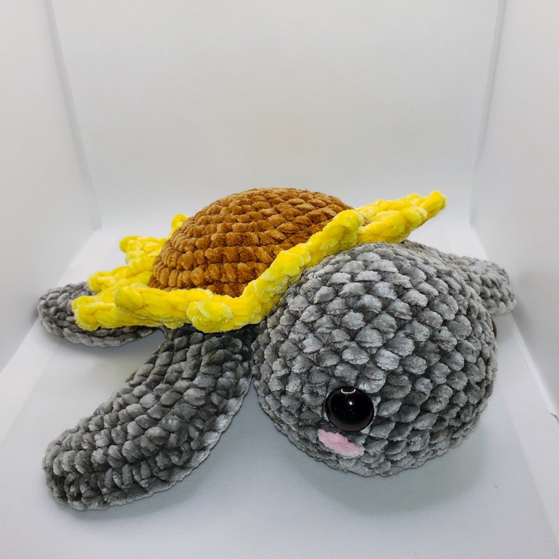 May include: A crocheted stuffed toy turtle with a yellow and brown sunflower on its back. The turtle has a gray body with a textured surface and a single black eye with a pink nose.
