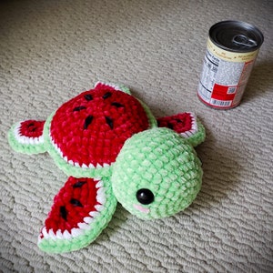 May include: A crocheted watermelon turtle plush toy with a green shell and red and white watermelon shell. The toy has a cute face with black eyes and a pink nose.