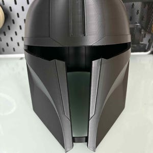 May include: A black Mandalorian helmet, 3D-printed with a matte finish. The helmet features a distinctive T-shaped visor, angled cheek plates, and a rounded top. The design is inspired by the Star Wars franchise.