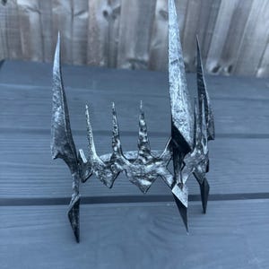 May include: A black metal crown with a pointed, jagged design. The crown is made of a textured metal and has a sharp, menacing look.