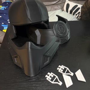 May include: A black, modular helmet with a rounded top and angular features. The helmet is composed of several pieces, including a face mask, side components, and a top section. Small white pieces are also present.
