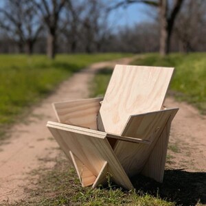 May include: A wooden chair with a unique, geometric design. The chair is made of plywood and has a natural wood finish. It is sitting on a dirt path with green grass on either side.