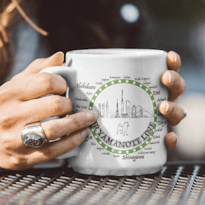May include: White ceramic mug with a green circle design featuring a cityscape and the text "Yamanote Line". The circle design includes the names of various train stations in Japanese.