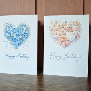 May include: Two blank greeting cards with a heart-shaped floral design. The card on the left features blue forget-me-not flowers and the card on the right features pink and white roses. Both cards say "Happy Birthday".