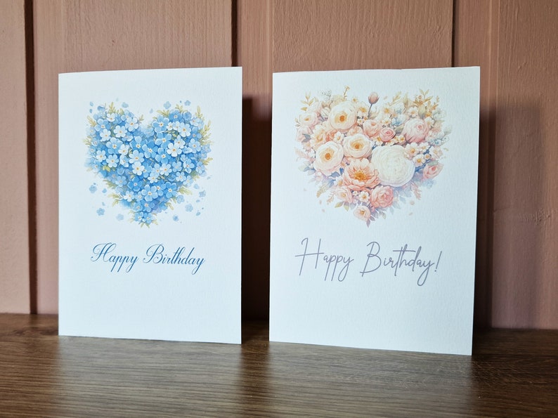 May include: Two blank greeting cards with a heart-shaped floral design. The card on the left features blue forget-me-not flowers and the card on the right features pink and white roses. Both cards say "Happy Birthday".