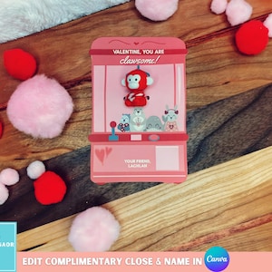 Könnte beinhalten: Eine rosa Valentinstagskarte in Form eines Greifautomaten mit dem Text "VALENTINE, YOU ARE clawsome!" und "YOUR FRIEND LACHLAN." Die Maschine enthält Plüschtiere. Rote und rosa Pompons sind um die Karte auf einer Holzoberfläche verstreut.