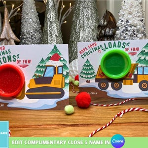 May include: Two Christmas cards with the text "Hope your Christmas is tons of fun!" and "Hope your Christmas is loads of fun!". One card features a red lid and a yellow bulldozer, the other a green lid and a yellow truck. The cards are on a wooden surface.
