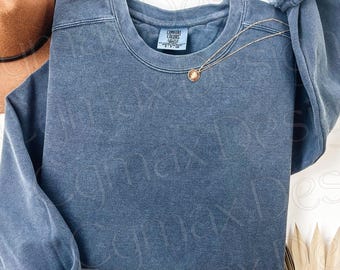May include: A blue jean colored crewneck sweatshirt with a Comfort Colors brand label at the inner neckline. The garment features long sleeves with ribbed cuffs and a solid, washed-out fabric texture. A thin gold chain necklace rests on the chest.
