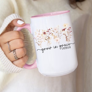 May include: White ceramic mug with a pink interior and handle. The mug features a floral design with the words "grow in grace" and the Bible verse 2 Peter 3:18 in black script. The mug is held by a person wearing a white sweater.