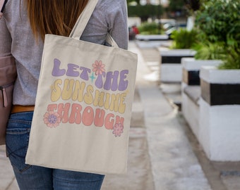 Peut inclure: Un sac fourre-tout blanc avec le texte "Let the Sunshine Through" dans une police rétro avec des fleurs colorées.