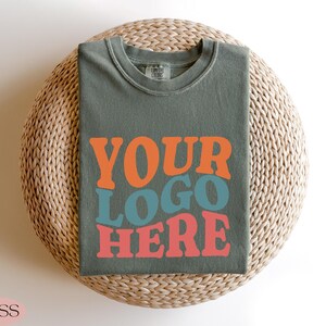 May include: A moss green t-shirt with the words "YOUR LOGO HERE" in orange, teal, and coral lettering. The shirt is folded and placed on a woven, circular surface. The word "MOSS" is in the bottom left corner.