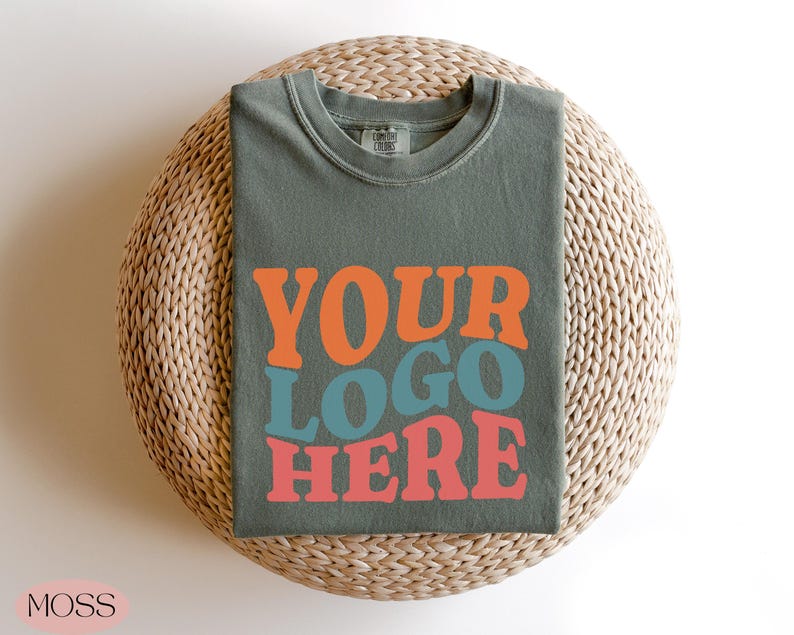 May include: A moss green t-shirt with the words "YOUR LOGO HERE" in orange, teal, and coral lettering. The shirt is folded and placed on a woven, circular surface. The word "MOSS" is in the bottom left corner.