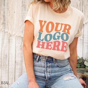 May include: Cream-colored t-shirt with the text "YOUR LOGO HERE" in orange, teal, and pink. The model is wearing blue jeans. The shirt has rolled-up sleeves and a crew neck. The word "NATURAL" is visible in the bottom left corner.
