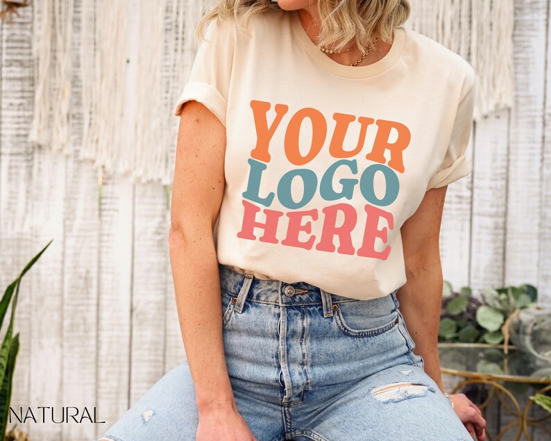 May include: Cream-colored t-shirt with the text "YOUR LOGO HERE" in orange, teal, and pink. The model is wearing blue jeans. The shirt has rolled-up sleeves and a crew neck. The word "NATURAL" is visible in the bottom left corner.