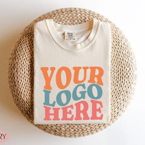 May include: Ivory-colored t-shirt with the text "YOUR LOGO HERE" in orange, blue, and pink. The shirt is folded and placed on a woven, circular surface. The word "IVORY" is in the bottom left corner.