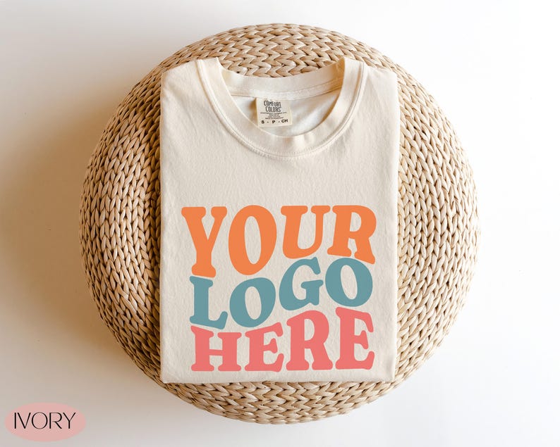 May include: Ivory-colored t-shirt with the text "YOUR LOGO HERE" in orange, blue, and pink. The shirt is folded and placed on a woven, circular surface. The word "IVORY" is in the bottom left corner.