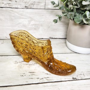 May include: Amber-colored glass shoe with a detailed, textured pattern. The vintage decorative piece has a small heel and a pointed toe. The shoe is displayed on a white wooden surface, with a potted plant in the background.