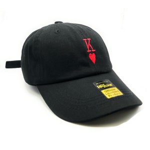 May include: Black baseball cap with a curved brim. The cap features red embroidered details of the letter "K" and a heart symbol. The cap has an adjustable strap and a "Hat Zone" official licensed product tag.