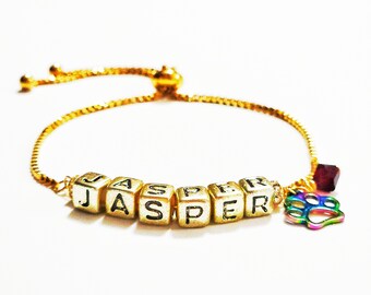 May include: Gold adjustable bracelet with a charm of a paw print and letter beads spelling out the name "JASPER".