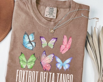 May include: A brown t-shirt with a faded look featuring a graphic of colorful butterflies and the text "FOXTROT DELTA TANGO".