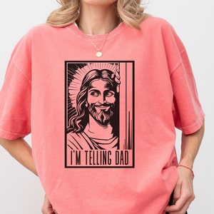 May include: A pink t-shirt featuring a black and white illustration of Jesus with the text "I'm telling Dad" underneath.