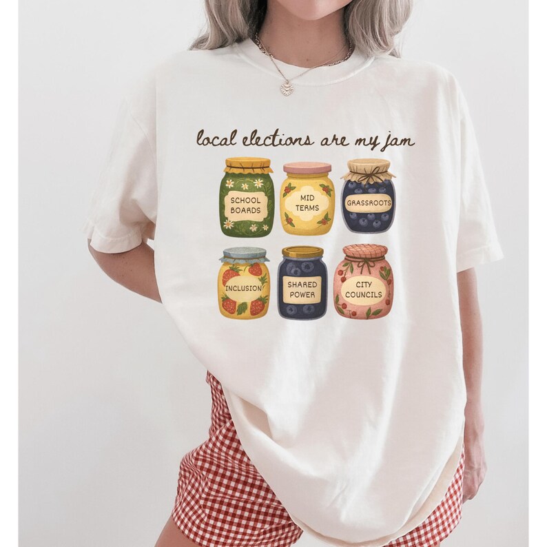 May include: Cream-colored t-shirt with the phrase "local elections are my jam" and illustrations of six jam jars. Each jar has a label with words like "School Boards" and "Inclusion". The shirt is a casual, everyday wear item.