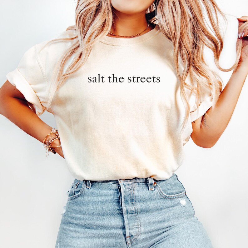 May include: Cream-colored t-shirt with the phrase "salt the streets" printed in black. The shirt is paired with blue denim jeans. The overall style is casual and relaxed.