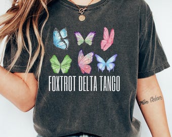 May include: A dark gray t-shirt with a distressed look featuring a graphic of six colorful butterflies and the text "FOXTROT DELTA TANGO".
