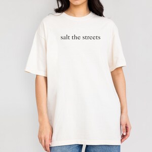 May include: Cream-colored t-shirt with the phrase "salt the streets" printed in black. The shirt has short sleeves and a relaxed fit. The text is centered on the chest.