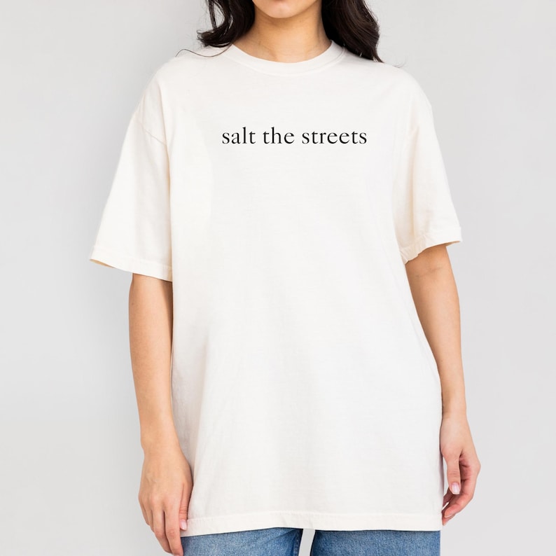 May include: Cream-colored t-shirt with the phrase "salt the streets" printed in black. The shirt has short sleeves and a relaxed fit. The text is centered on the chest.