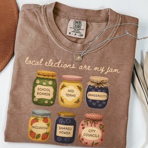 May include: A brown Comfort Colors t-shirt with the phrase "local elections are my jam." The shirt features illustrations of six jars labeled "School Boards," "Mid Terms," "Grassroots," "Inclusion," "Shared Power," and "City Councils."