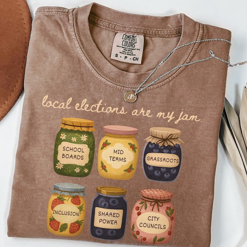 May include: A brown Comfort Colors t-shirt with the phrase "local elections are my jam." The shirt features illustrations of six jars labeled "School Boards," "Mid Terms," "Grassroots," "Inclusion," "Shared Power," and "City Councils."